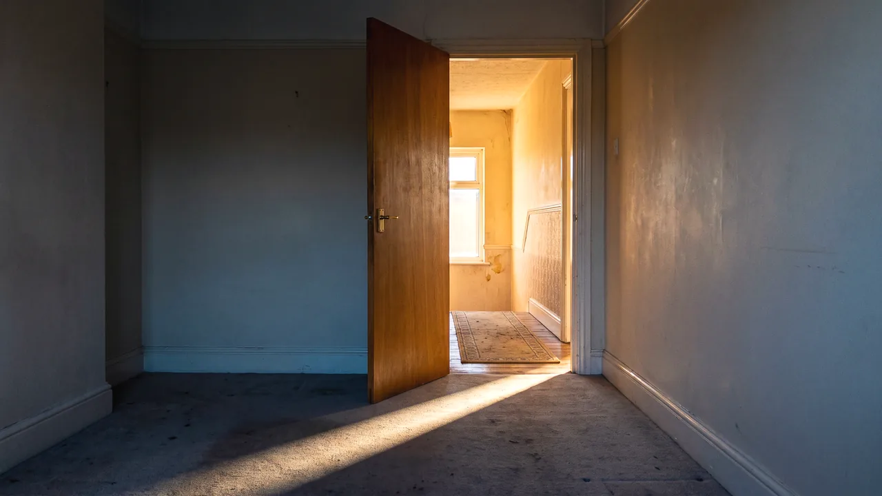 A door standing open onto a bright landing, the near room dim and cool, a hard bar of daylight thrown across the floor through the doorway.